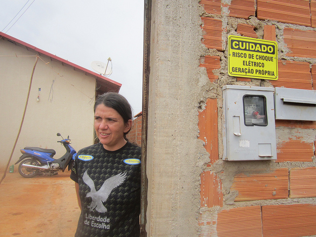 Patricia Soares de Oliveira, who was the first to receive solar panels as a test in 2017, stands in front of her house and next to an electric meter that reads &amp;quot;danger of electric shock&amp;quot;. Her power bill in this social housing complex on the outskirts of Palmeiras de Goiás in central Brazil has fallen to one-fifth of what she previously paid. Credit: Mario Osava/IPS