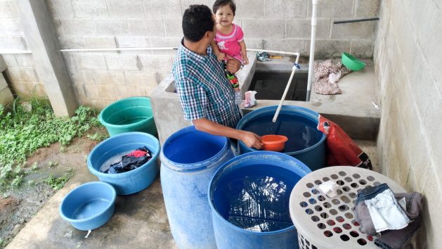 Alex Leiva, holding his baby girl, uses the water he managed to collect in barrels at 4:00 a.m., the only time the service is provided in Lotificación Praderas, in the canton of Cabañas, on the outskirts of the municipality of Apopa, north of the Salvadoran capital. The families of this region are fighting in defense of water, against an urban development project for wealthy families that threatens the water resources in the area. CREDIT: Edgardo Ayala/IPS