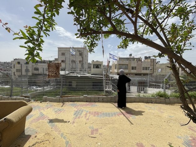72-year-old Kawthar Ajlouni stands alone in her yard in H2, Hebron, the occupied Palestinian territory. The backdrop reveals a fortified Israeli checkpoint. Amid 645 documented movement obstacles in the West Bank, 80 are here in H2 as of 2023. Isolated due to strict Israeli policies, she is one of 7,000 Palestinians enduring heavy restrictions, while many others have left. The Israeli-declared 'principle of separation' (between Palestinians and Israeli settlers) limits their life, generating a coercive environment that risks forcible transfers. Kawthar stays, fearing her home's conversion into a military post. Credit: OCHA/2023