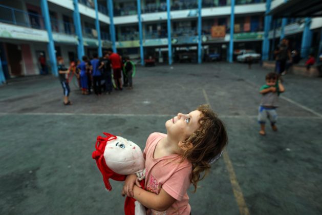 Aya, a 5-year-old girl clutching her doll to ease her fear, gazes at Gaza's sky filled with warplanes from inside an UNRWA school in the Gaza Strip. Credit: UNRWA